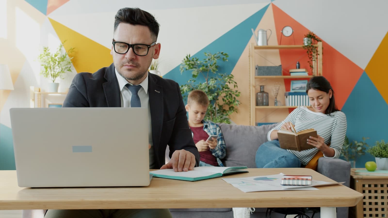 Business owner in suit working at desk with laptop, family visible in background