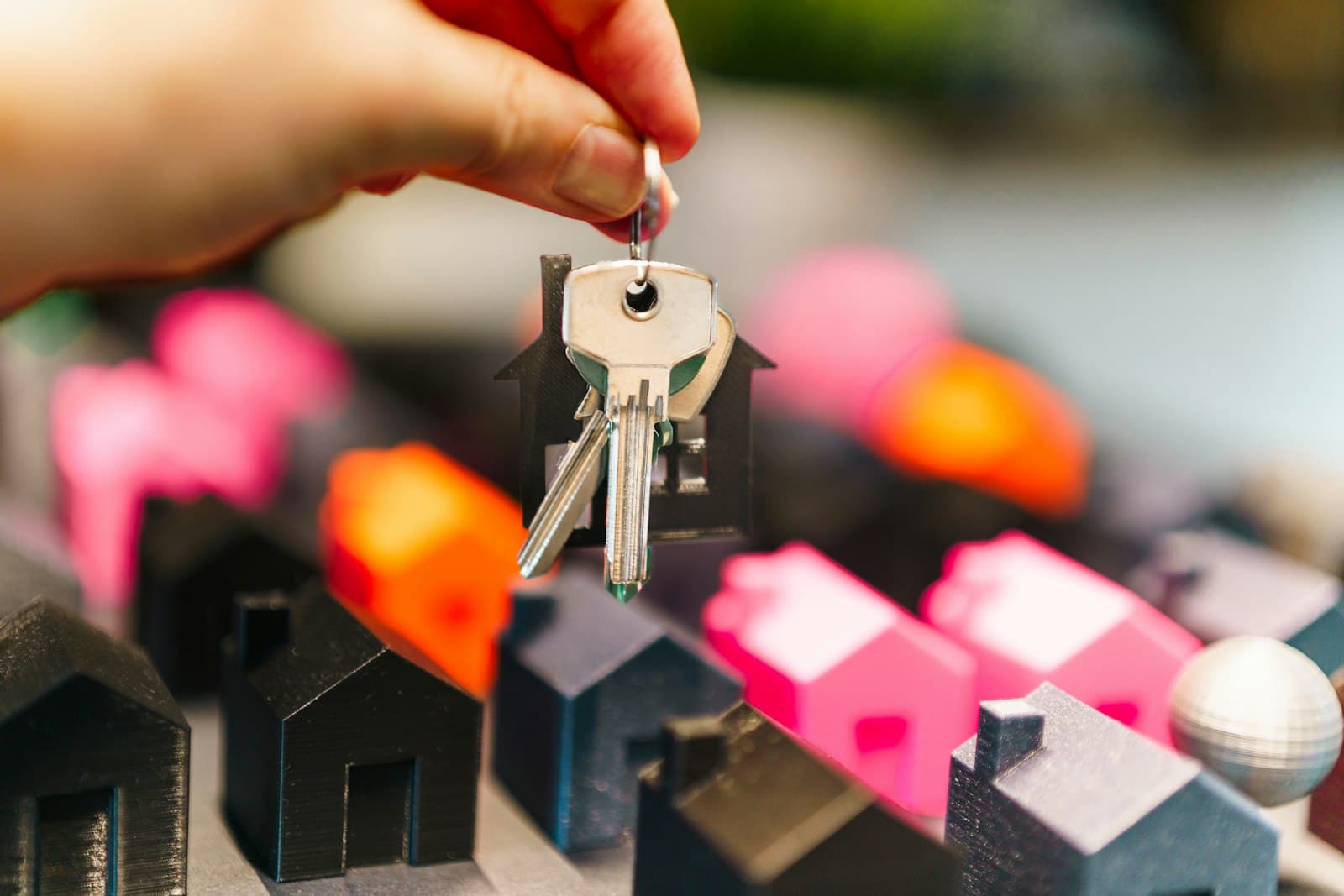 Hand holding a house key with a house-shaped keychain above colorful miniature model houses — property transfer