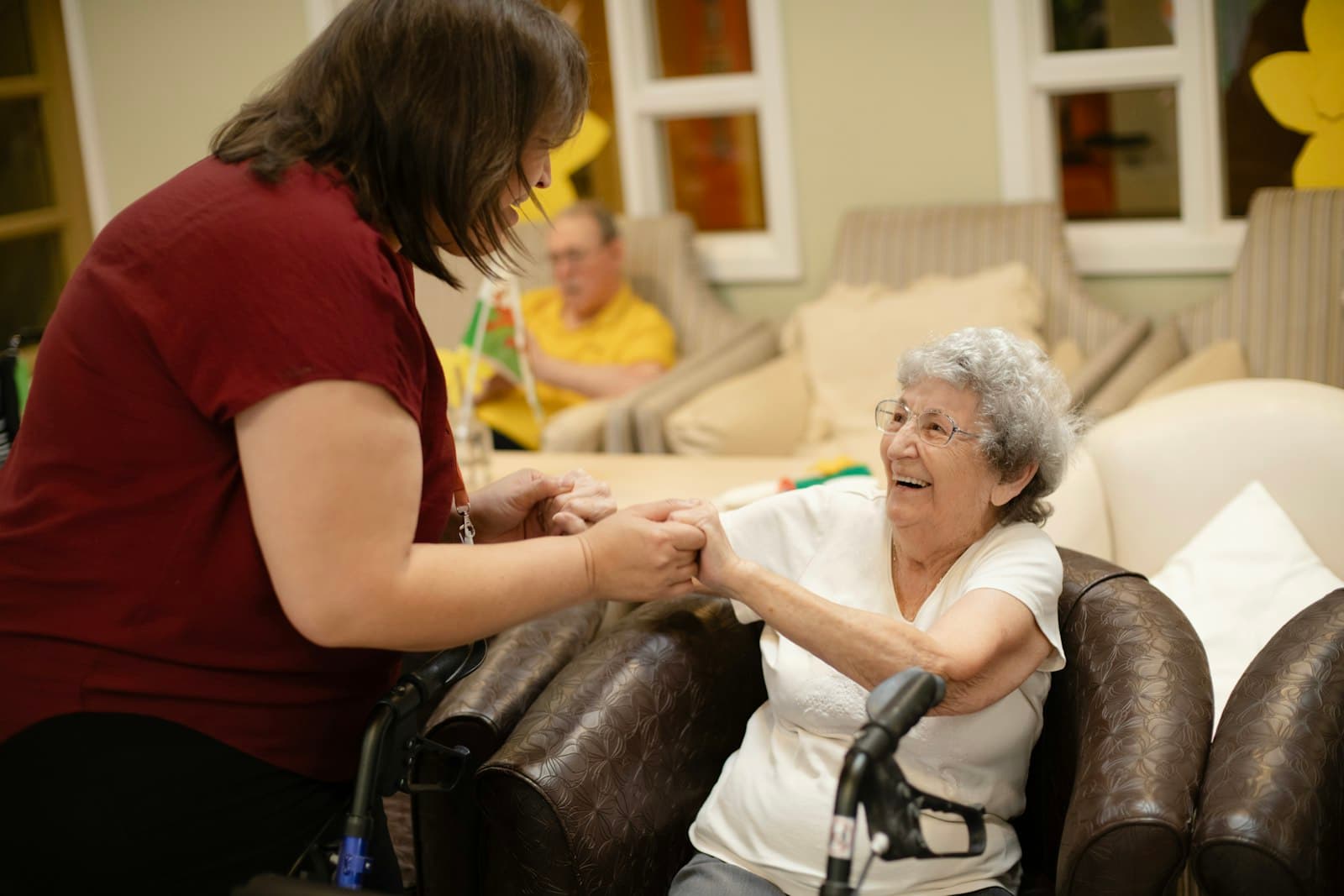 Caregiver in red shirt warmly holding hands with an elderly woman seated in a care facility armchair