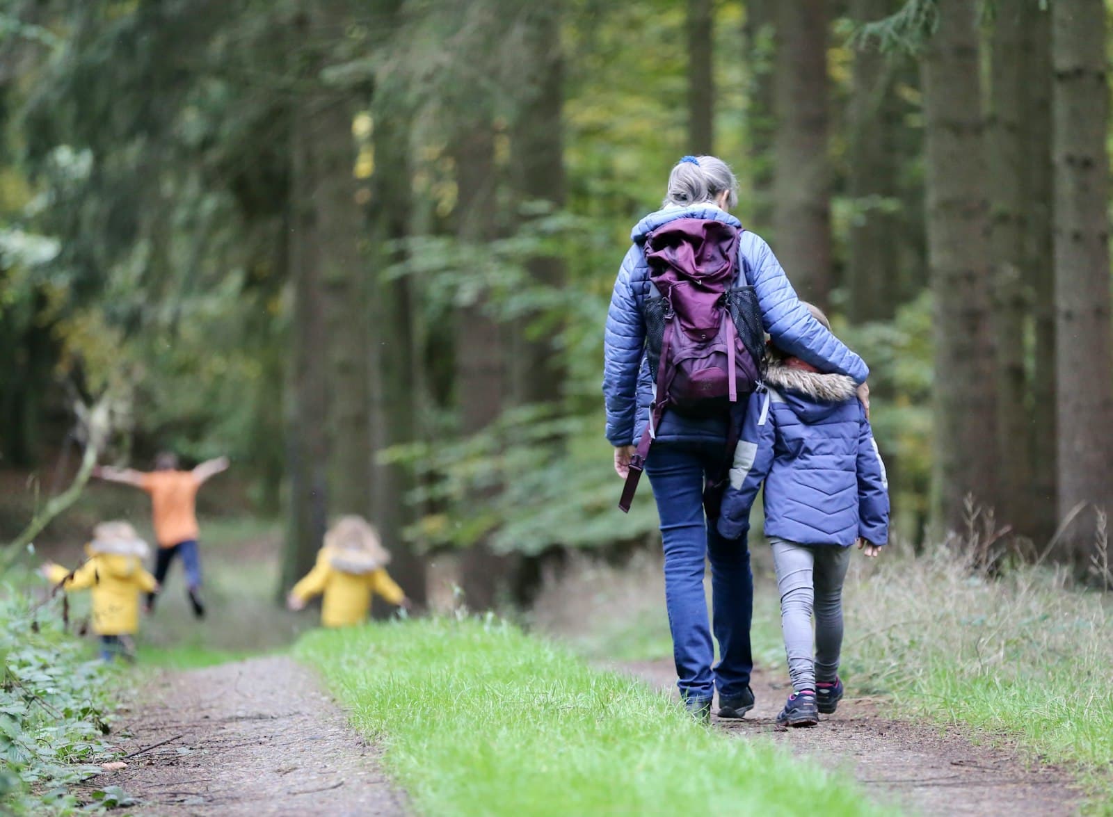 Grandmother walking with grandchildren along a tree-lined forest path — generational family legacy