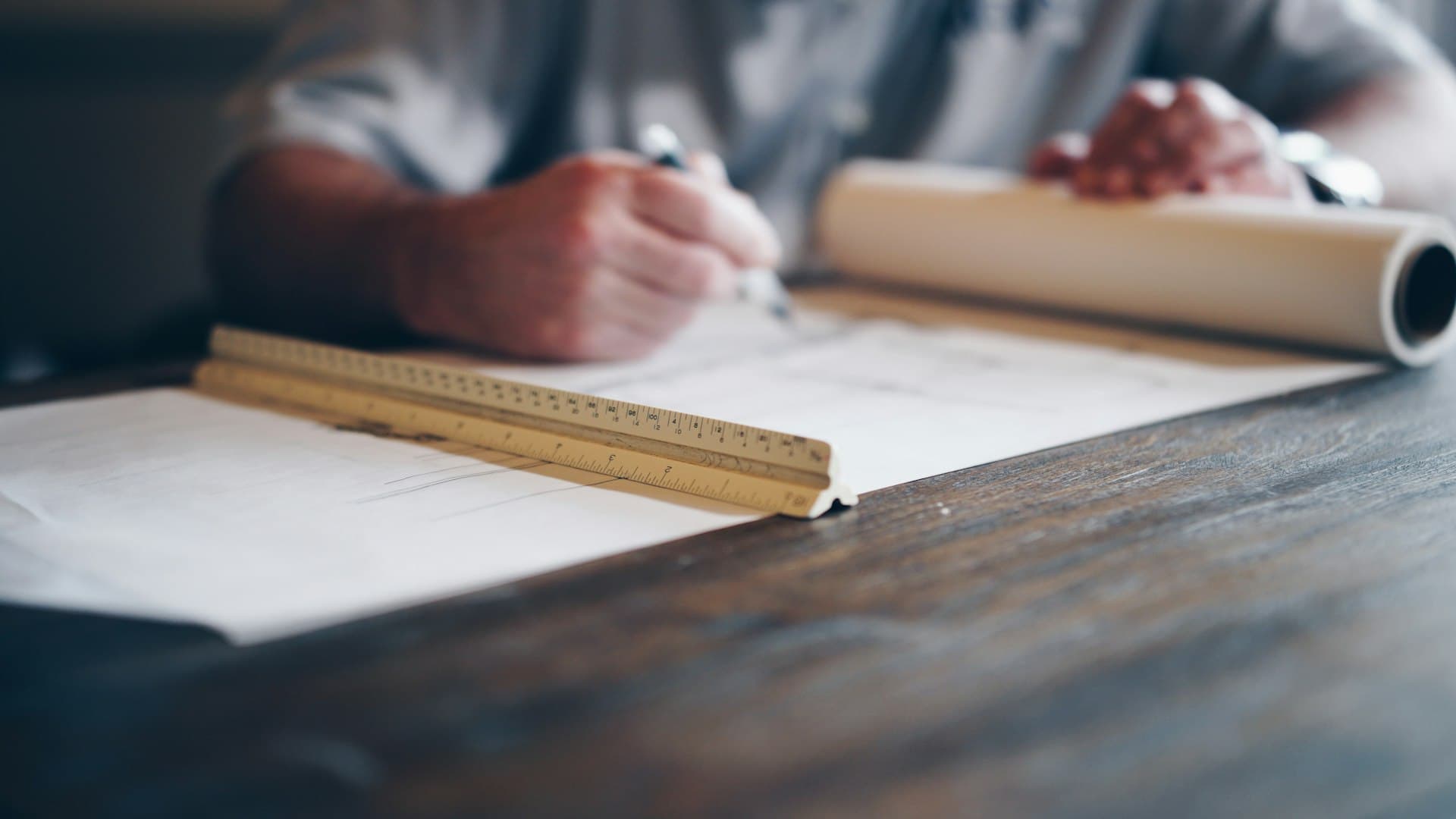 Hands drawing on an architectural plan with a scale ruler on a dark wooden desk