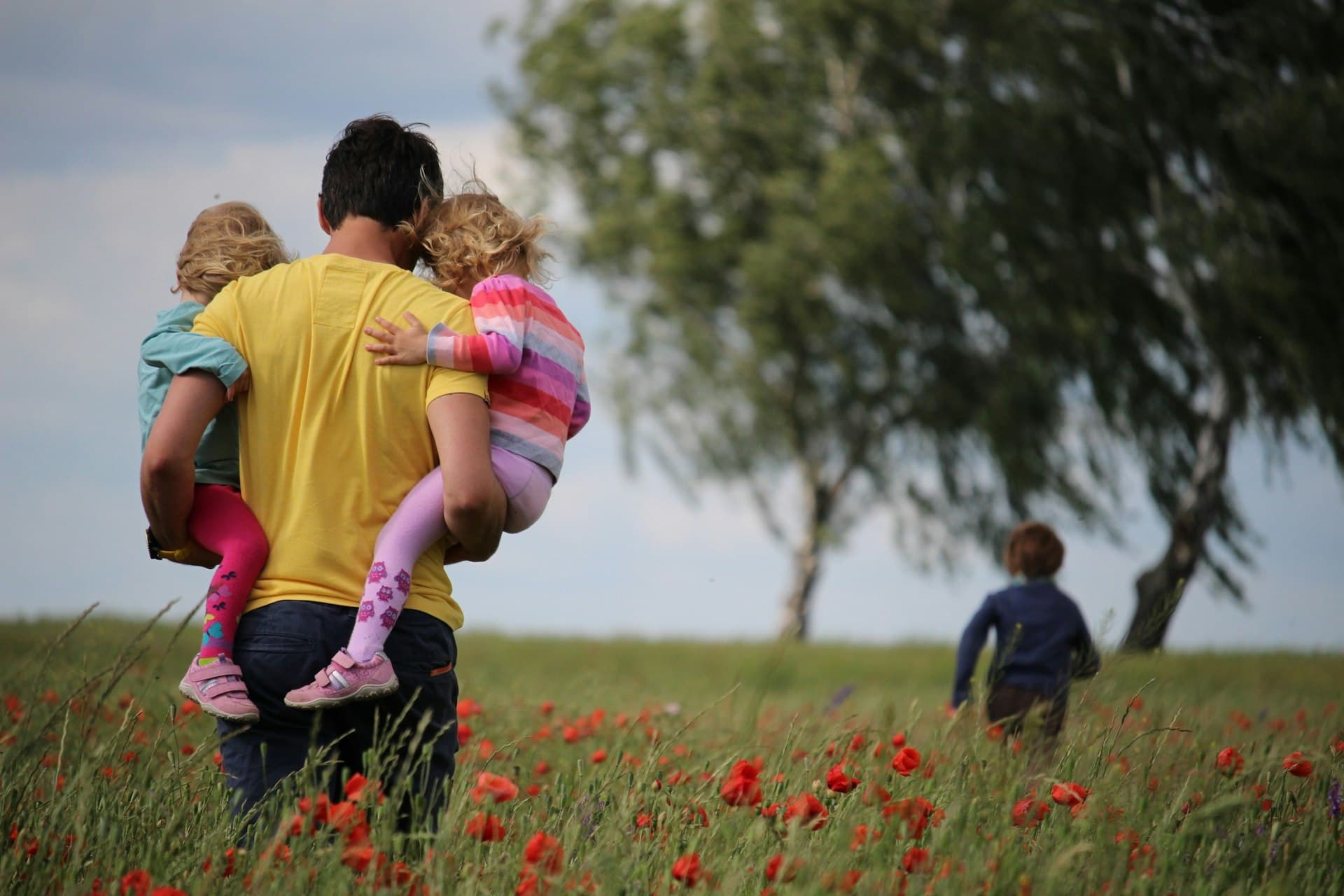 Father carrying two young children through a sunlit field of red flowers, a third child running ahead