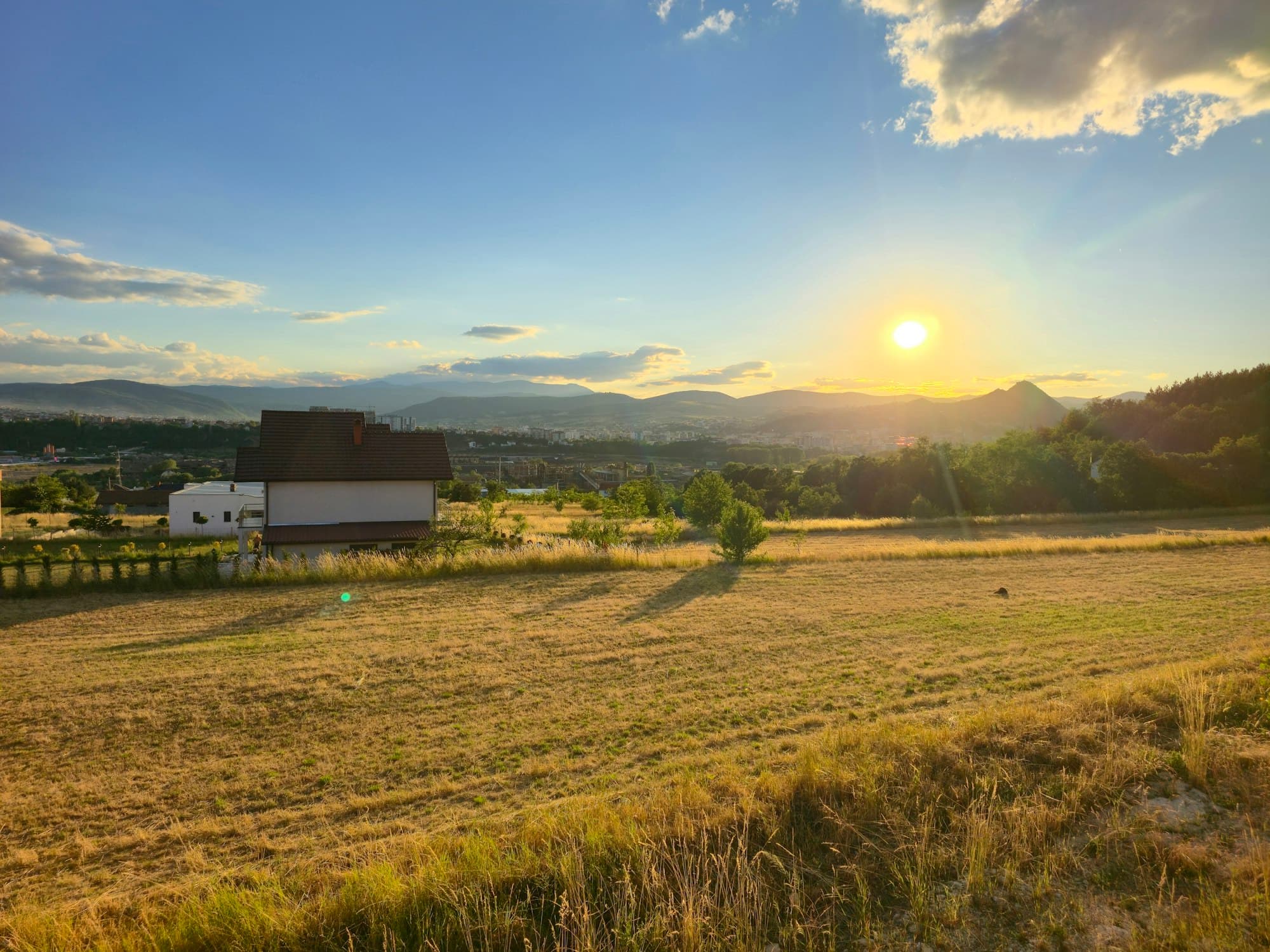 White farmhouse set in a golden meadow at sunset with rolling hills in the background